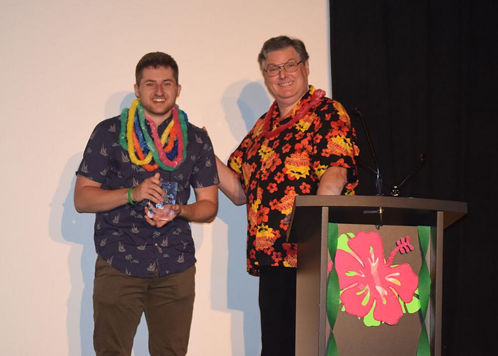 Two men in Hawaiian shirts and leis smile next to a podium.