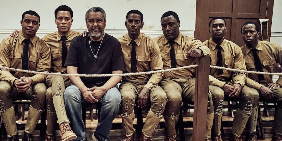 A row of several men sit on a bench in a courtroom film set.