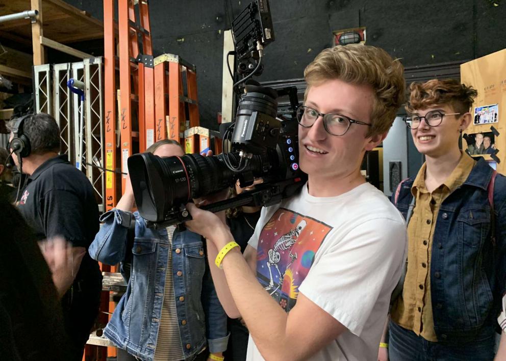 A student in a T-shirt holds a camera backstage as other students look on.