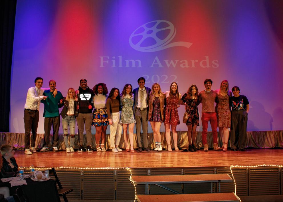 A group of students stand with their arms around each other on a stage in front of a film reel logo that reads, "Film Awards 2022"