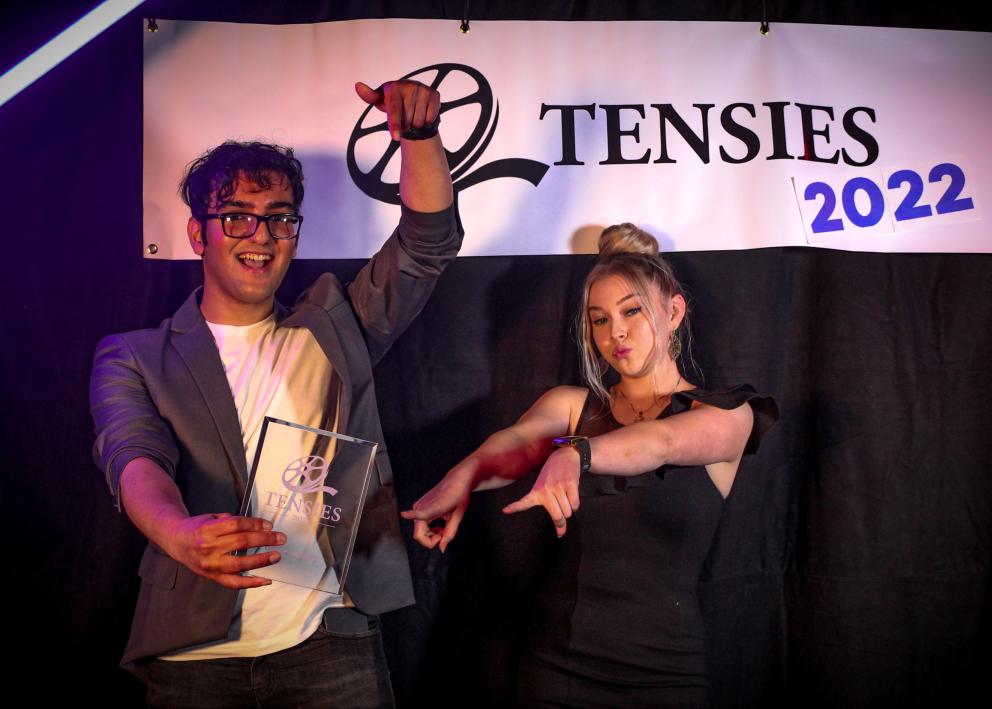 Two students stand in front of a Tensies 2022 banner, holding up a clear acrylic Tensies trophy.