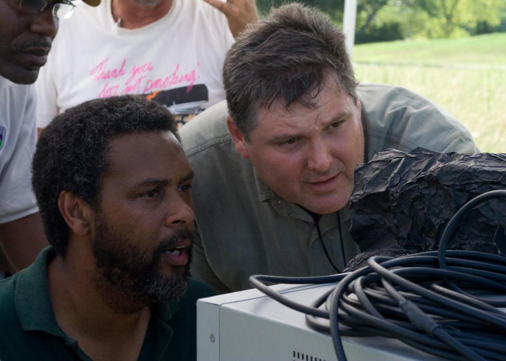 Two men look through a film camera with a film crew standing behind them.