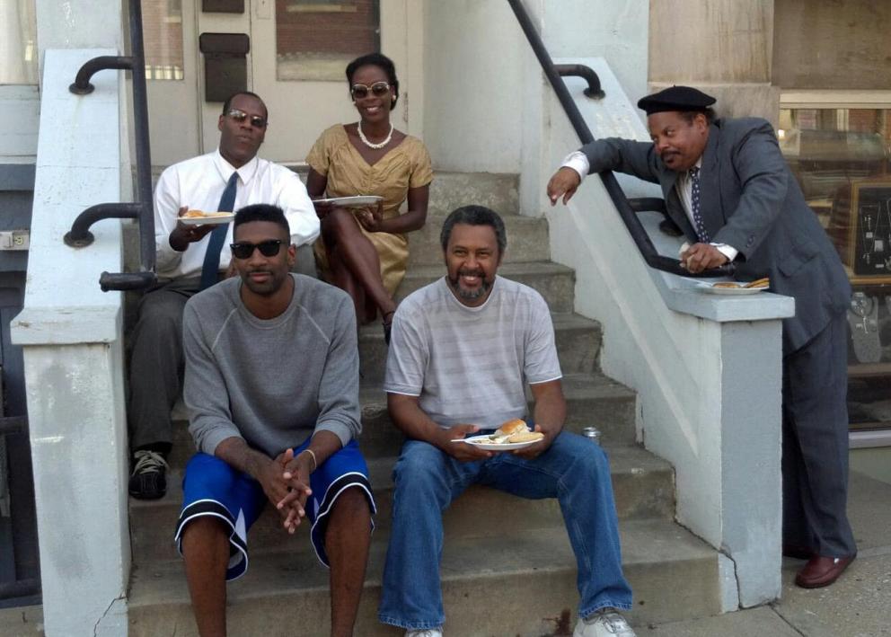 Four people sit on cement steps with plates of food. Another man leans on the railing.