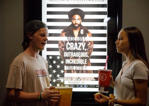 A boy and a girl chat in front of a "BlacKkKlansman" movie poster.