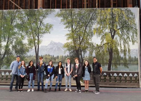 A group of students stand in front of a painted scene of a trees in front of a mountain.