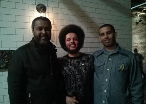 Three men stand in a hallway and smile for the camera.