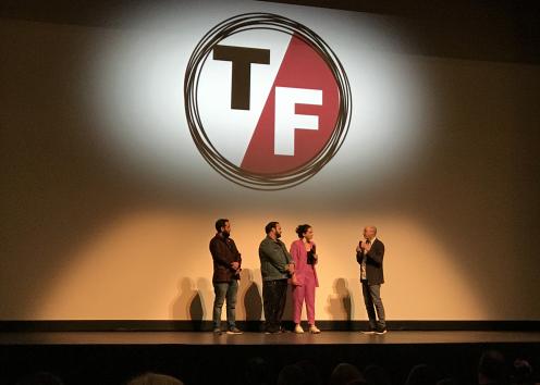 A man with a microphone talks to three other people on a stage with the True False Film Festival logo on a screen above them.